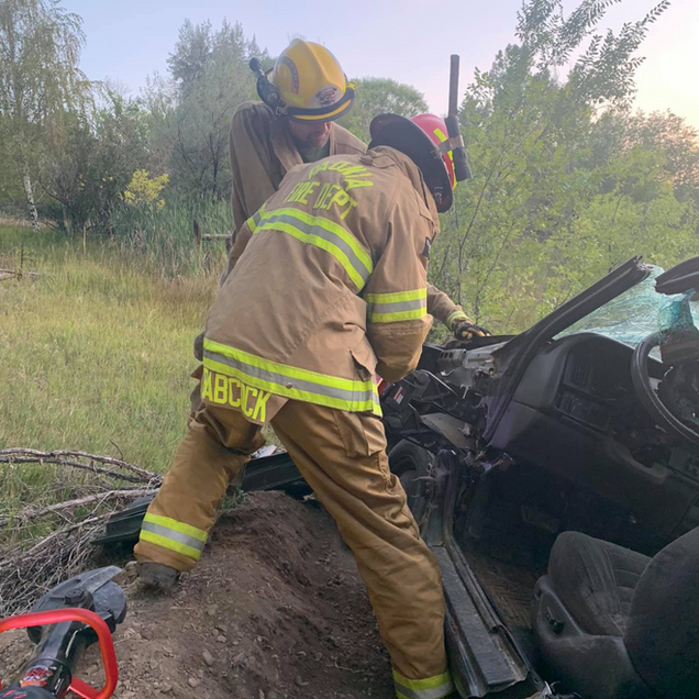 Firefighters at car crash scene; one inspects damage, another looks on.