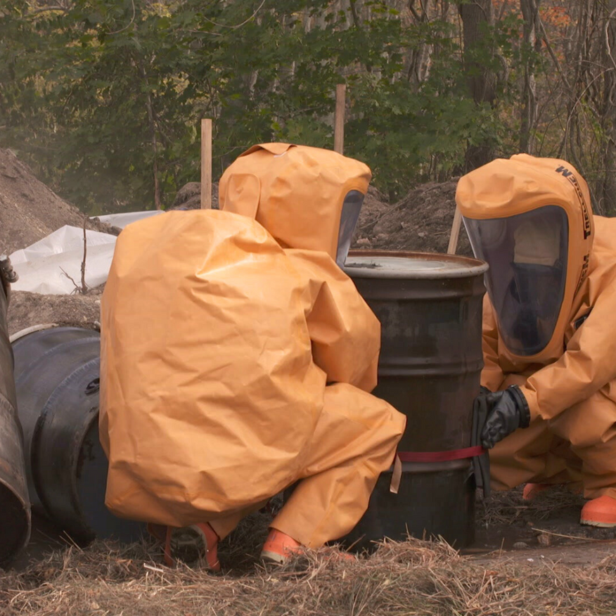 Three people in orange hazmat suits working near barrels outdoors.