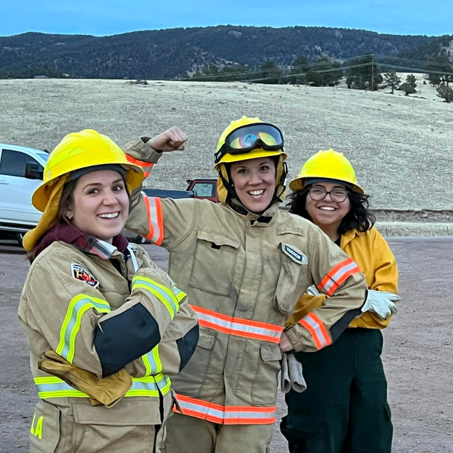 Three women in firefighting gear pose, one flexes arm. Yellow helmets and tan uniforms, outdoor setting.