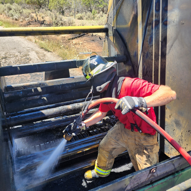 Firefighter spraying water inside charred machinery. Outdoor setting, red shirt, helmet, hose.