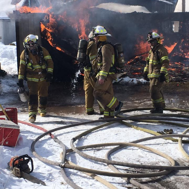 Firefighters battling a blaze at a structure; hoses and a chainsaw are in the foreground, snow on the ground.