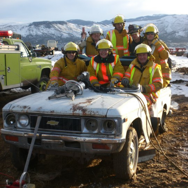Firefighters in safety gear practice vehicle extraction on a damaged pickup truck outdoors, mountains in background.