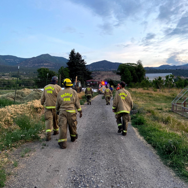 Firefighters in gear walk down a dirt road toward a structure with smoke billowing in the distance.