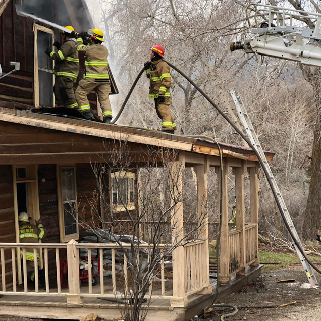 Firefighters on a roof and porch, battling a house fire with hoses and ladder, outdoors.