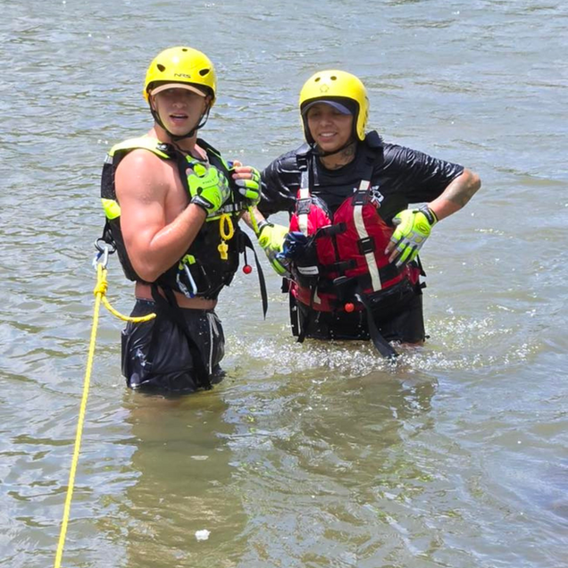 Two people in yellow helmets and life vests stand in water, holding ropes.