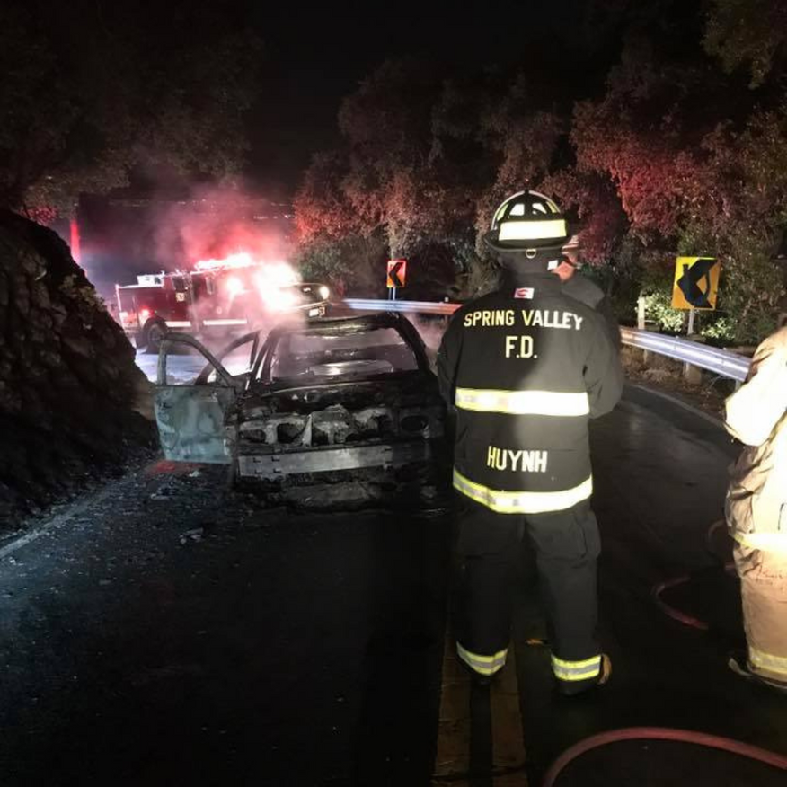 Firefighter Nguyen of Spring Valley F.D. stands on a dark road next to a burning car; fire trucks in the background.