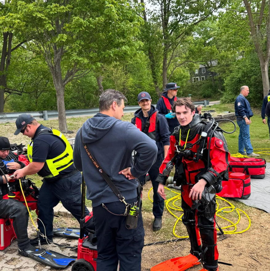 Divers and emergency personnel prepare for a water rescue near a shoreline; one diver in red suit.