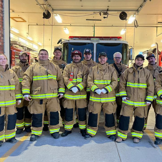 Firefighters in protective gear stand in front of a fire truck at a station.