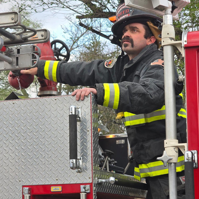 Firefighter in a black jacket, adjusting equipment on a red fire truck, wearing a helmet.