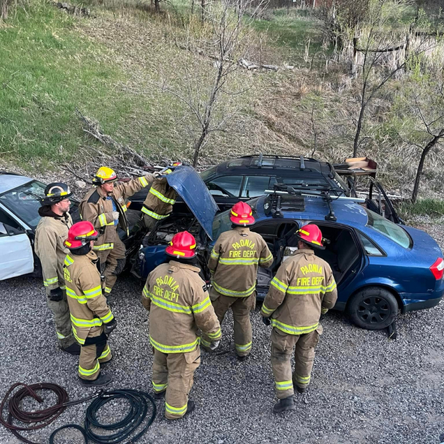 Firefighters surround a blue car; they are doing training exercises, preparing for extraction in a gravel lot.