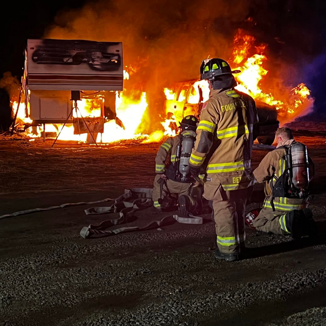 Firefighters battling a burning RV at night; flames and smoke are visible.