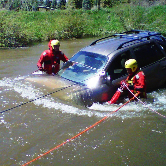 Two rescuers in water, one in vehicle, pulling submerged car from a river using ropes.