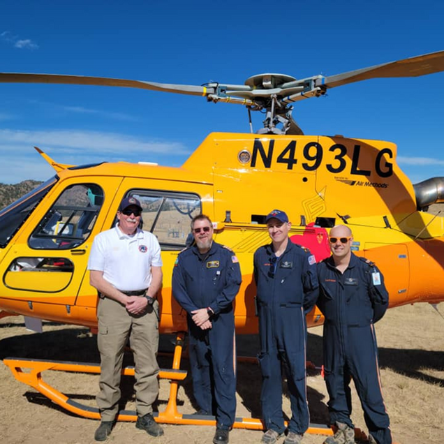 Four people stand in front of a bright yellow helicopter. The sky is blue.