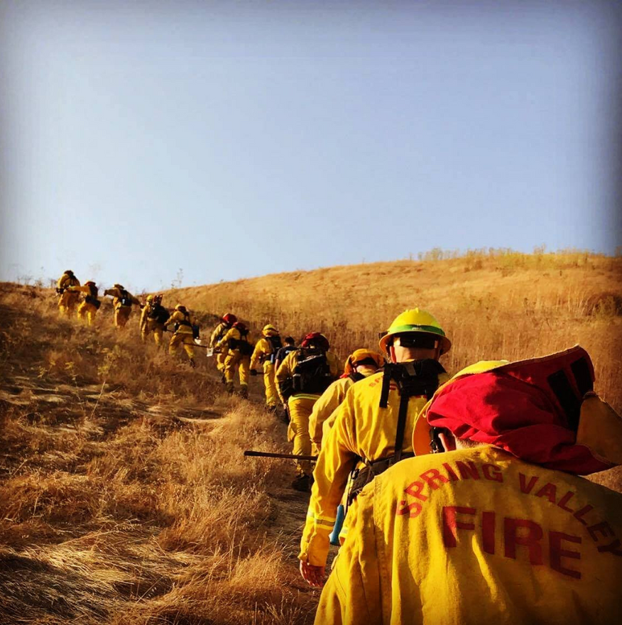Firefighters in yellow gear hiking up a dry, grassy hillside under a blue sky.