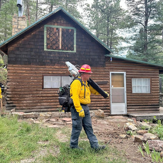 Firefighter in yellow gear, with equipment, standing near a wooden cabin in a forest.