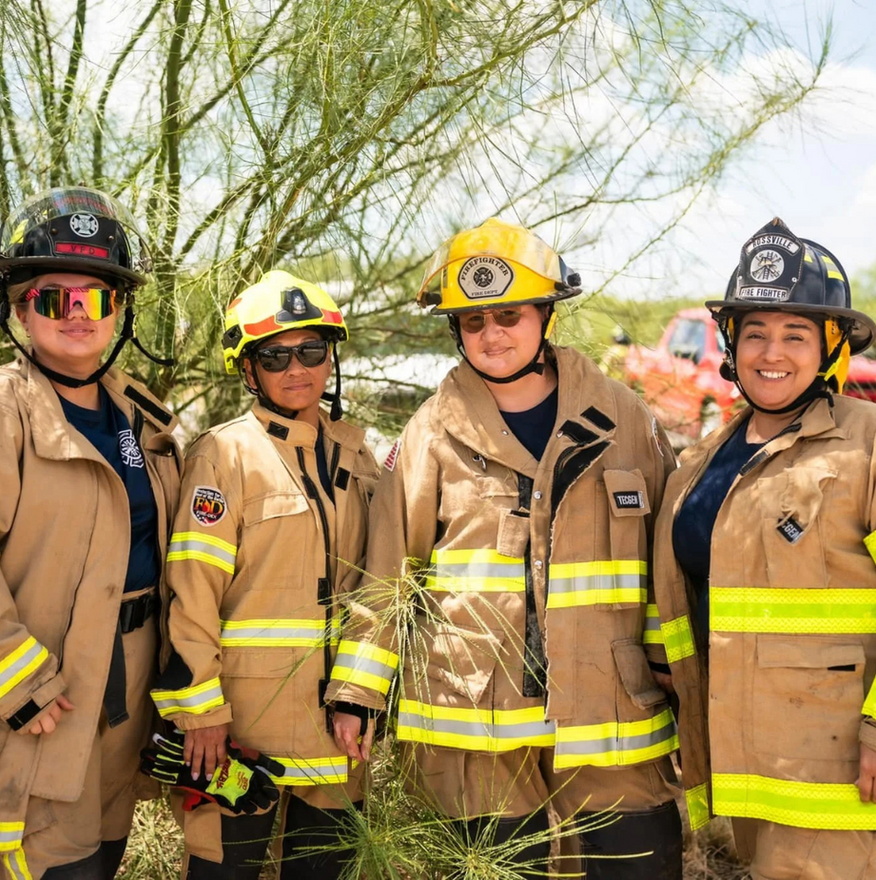 Four firefighters in tan coats and helmets stand outdoors.