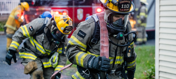 Firefighters wearing protective gear, entering a building, red fire truck in the background.