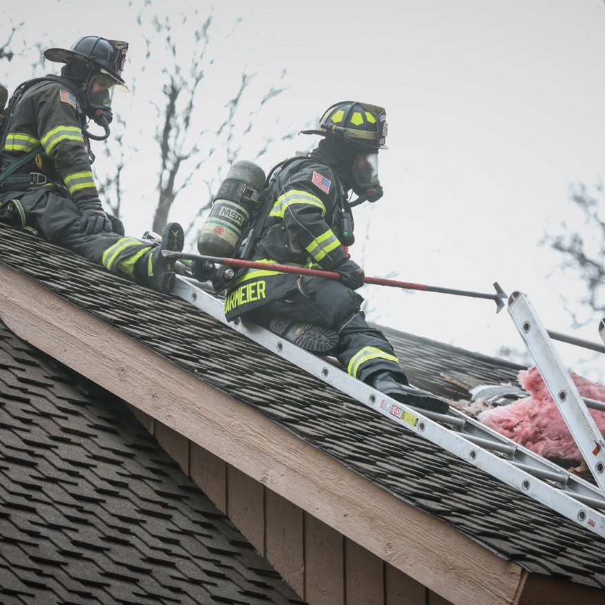 Firefighters on a rooftop, wearing masks, using a ladder; smoke and pink insulation visible.