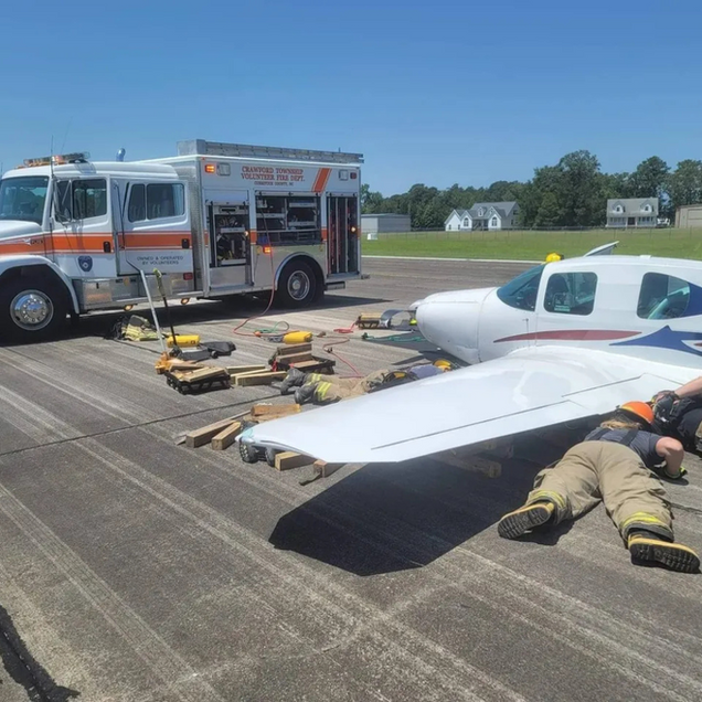 Plane on runway with emergency vehicle. Firefighter lies near wing; supports and equipment surround.