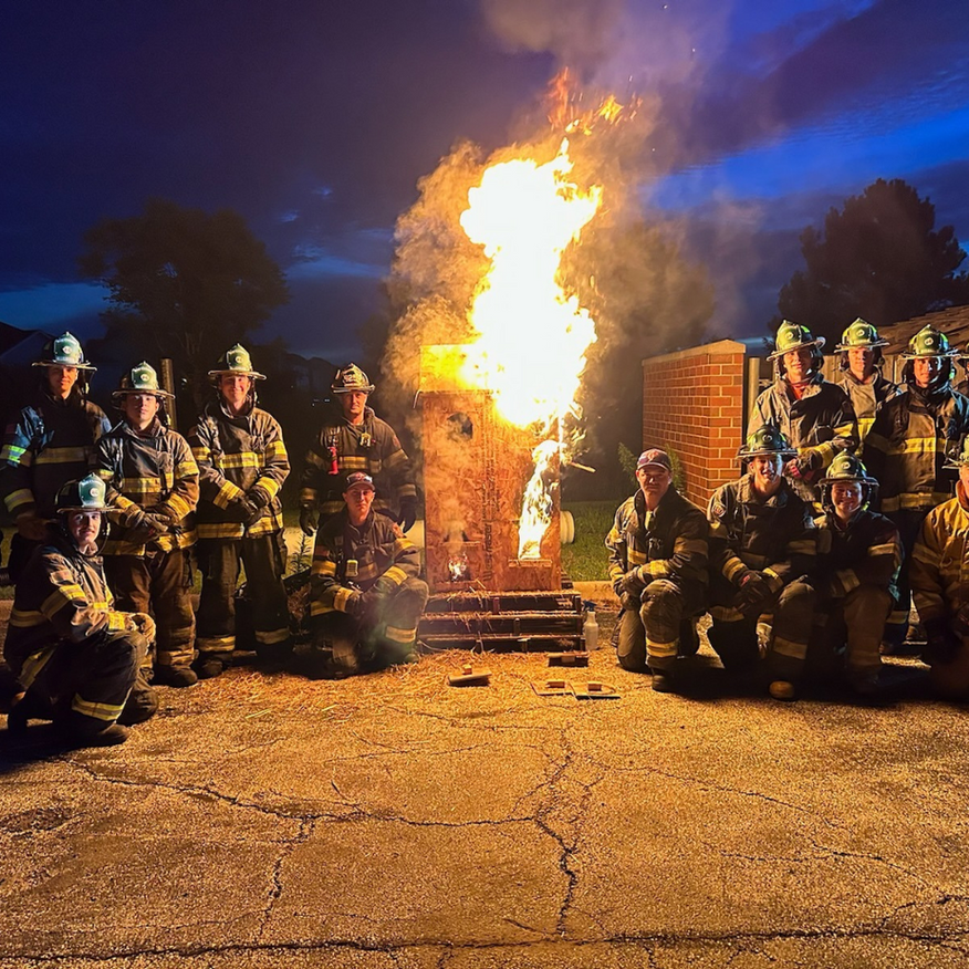 Firefighters in uniform pose in front of a burning structure at night.