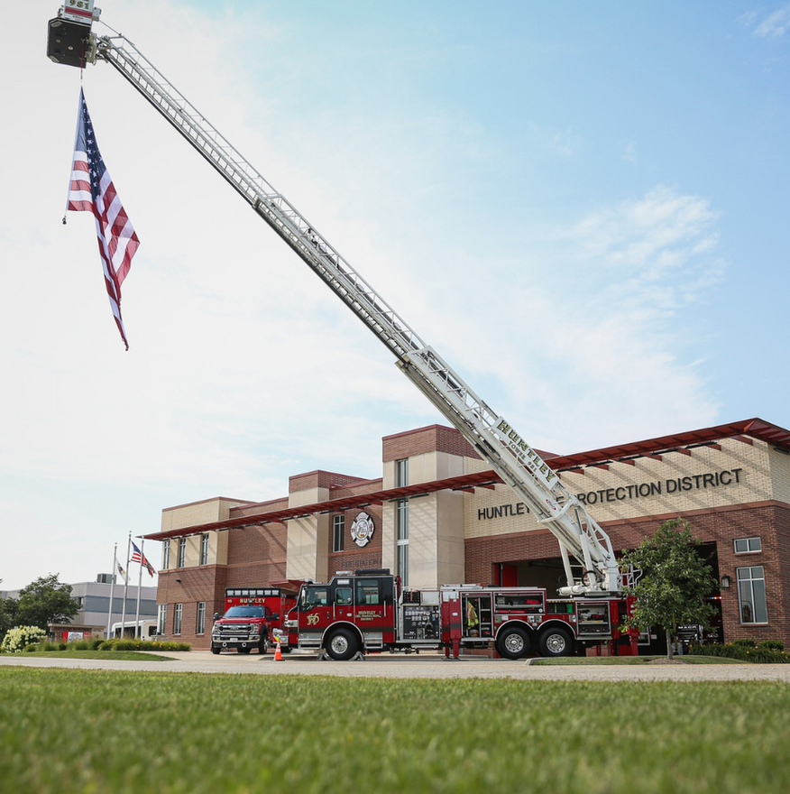 Fire truck with extended ladder holding an American flag in front of a fire station.