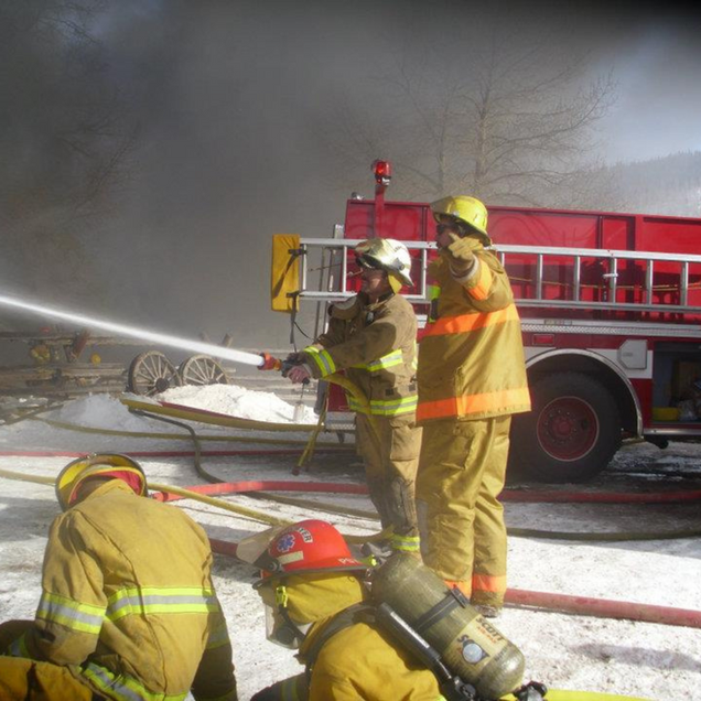 Firefighters in yellow gear battle a fire, spraying water near a red fire truck. Smoke billows in the background.