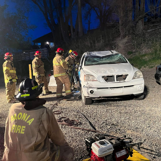 Firefighters at a vehicle accident at dusk, assessing damage and using rescue equipment.