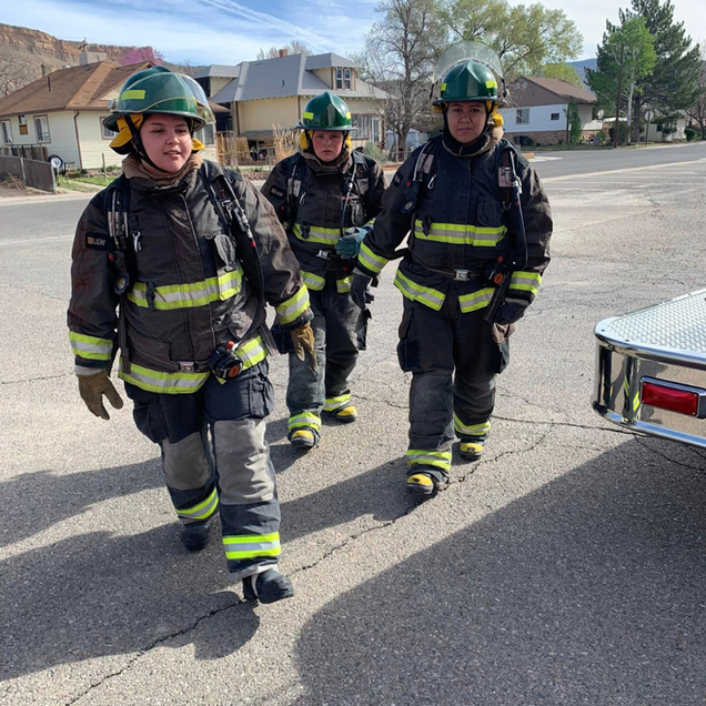 Three firefighters in full gear walk on a street near buildings and a fire truck.