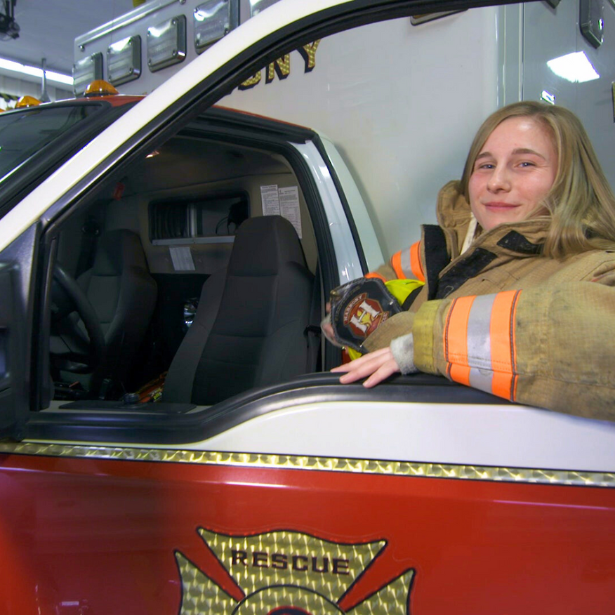 Female firefighter in uniform, holding helmet, seated in a rescue truck.