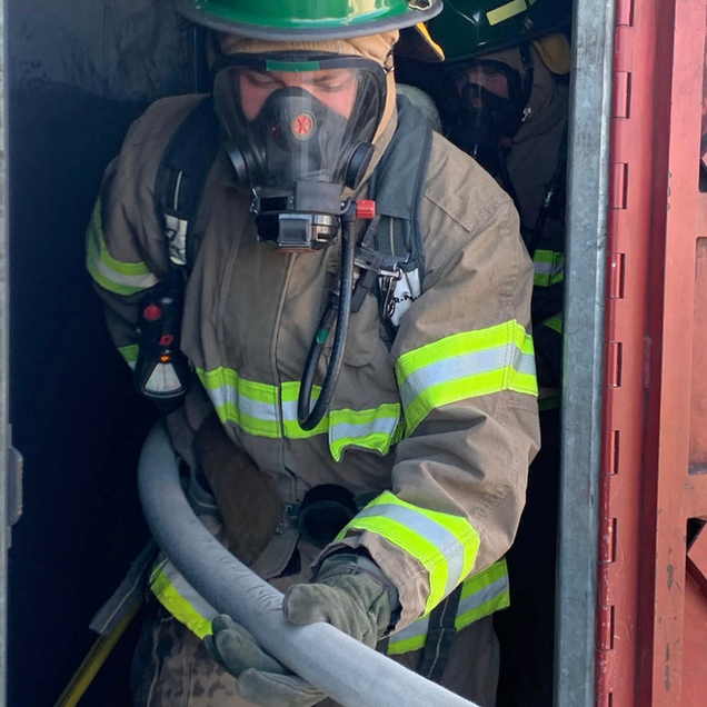 Firefighter in full gear, holding a hose, emerges from a doorway.