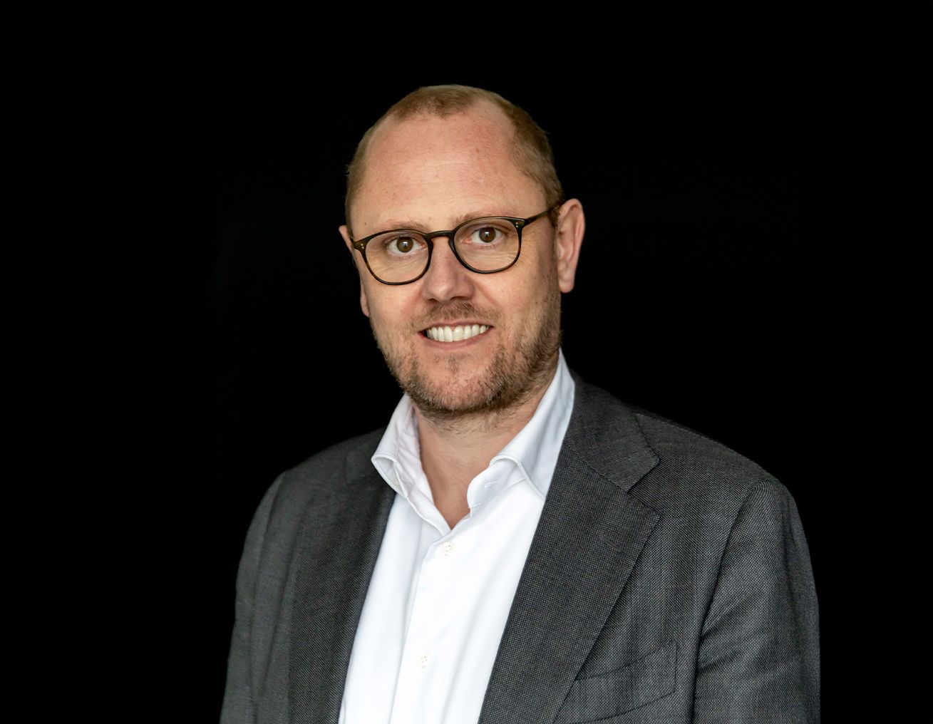 Een man met een bril en een pak lacht naar de camera.