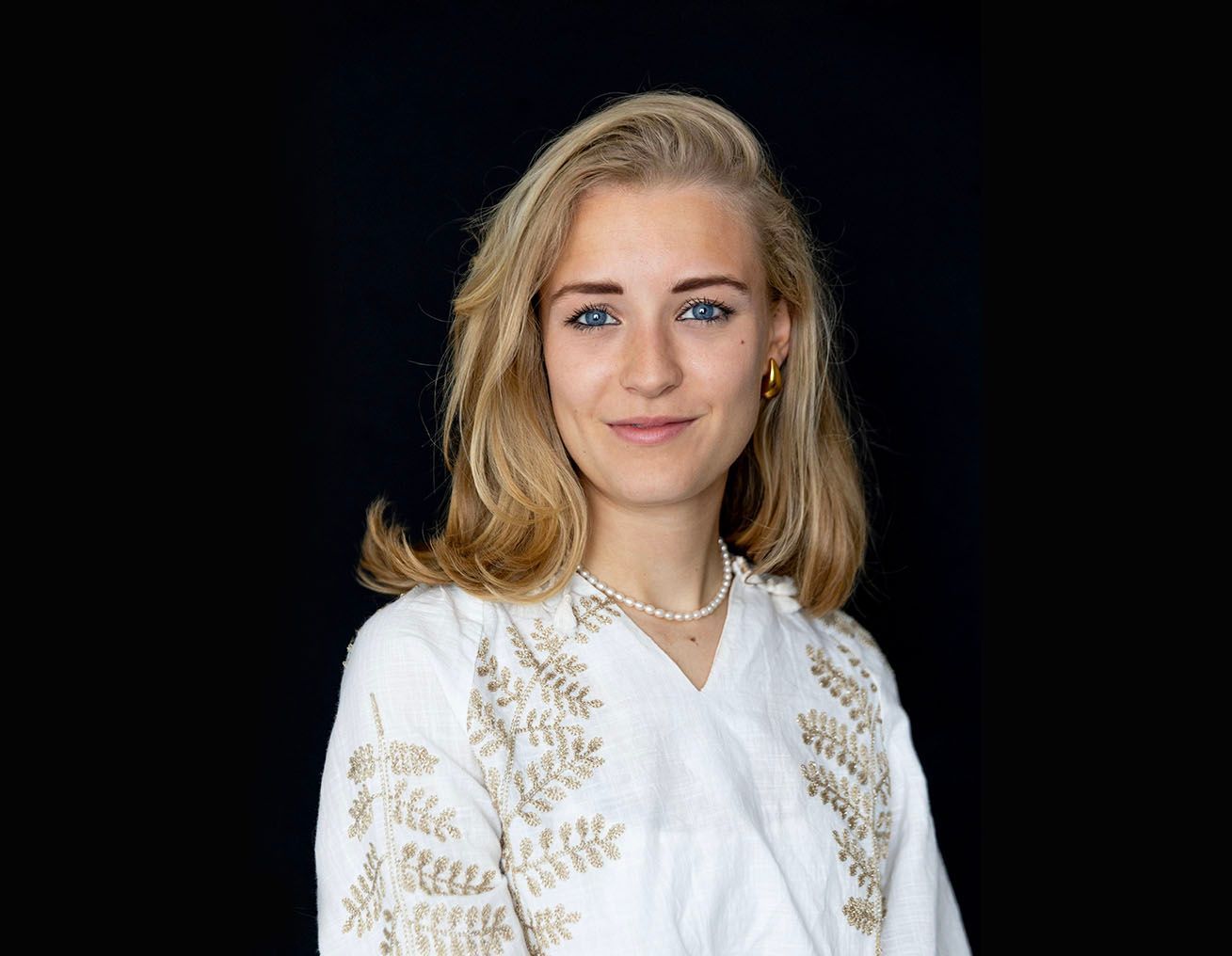 Een jonge vrouw, gekleed in een wit overhemd en een parelketting, staat voor een zwarte achtergrond.
