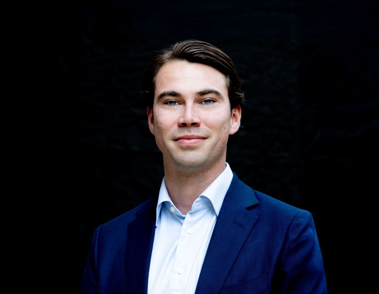Een man in een blauw pak en wit overhemd staat voor een zwarte achtergrond.