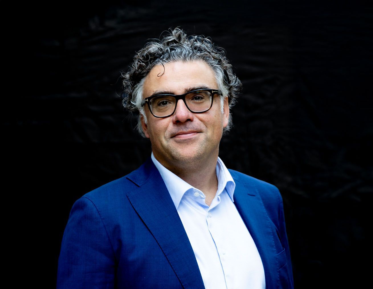 Een man met een bril en een blauw pak staat voor een zwarte achtergrond.