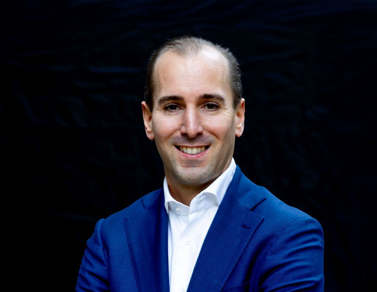 Een man in een blauw pak en wit overhemd lacht naar de camera.