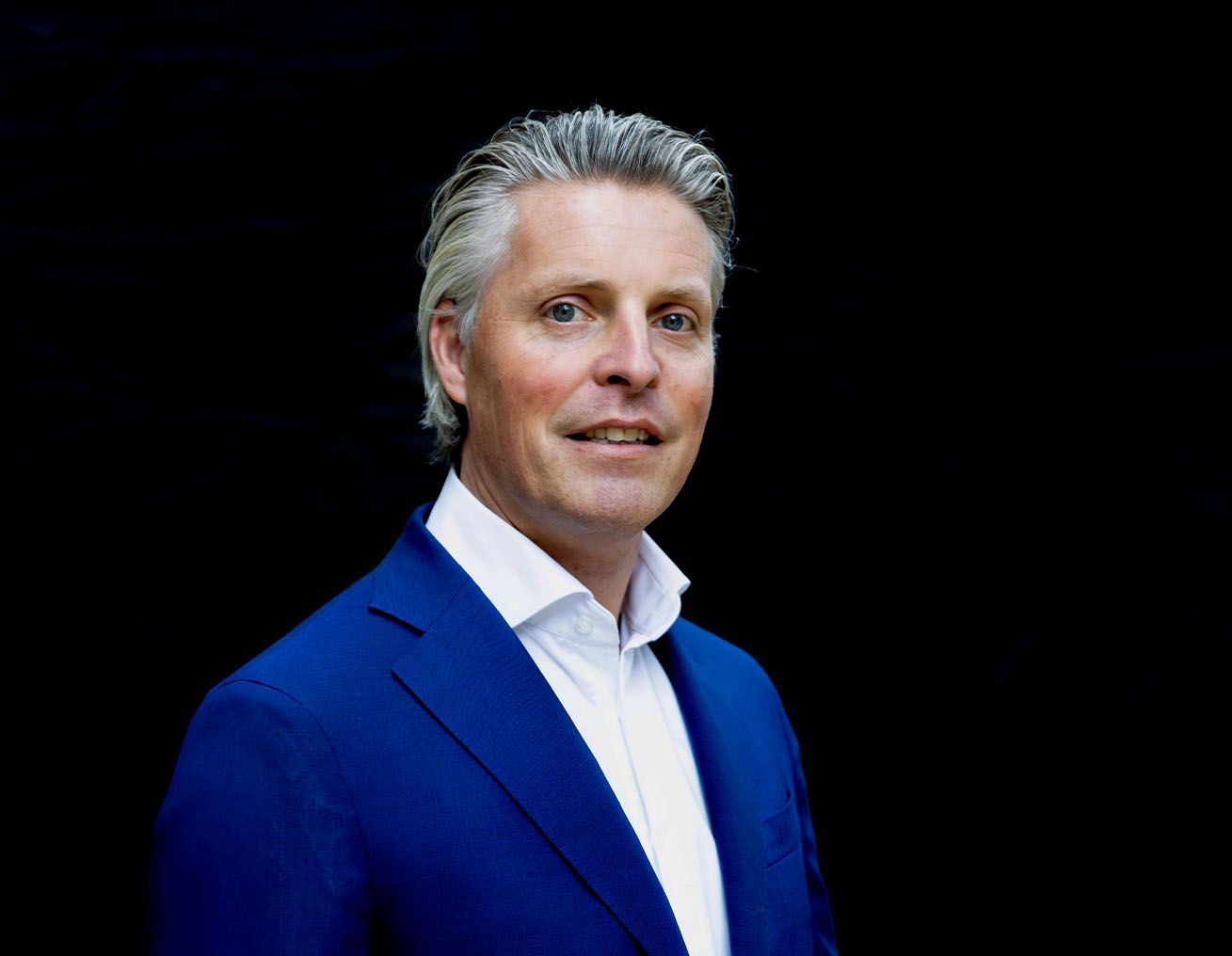 Een man in een blauw pak en wit overhemd staat voor een zwarte achtergrond.
