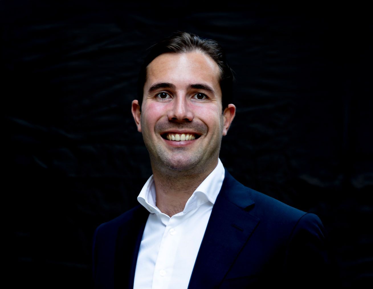 Een man in een pak en een wit overhemd lacht naar de camera.