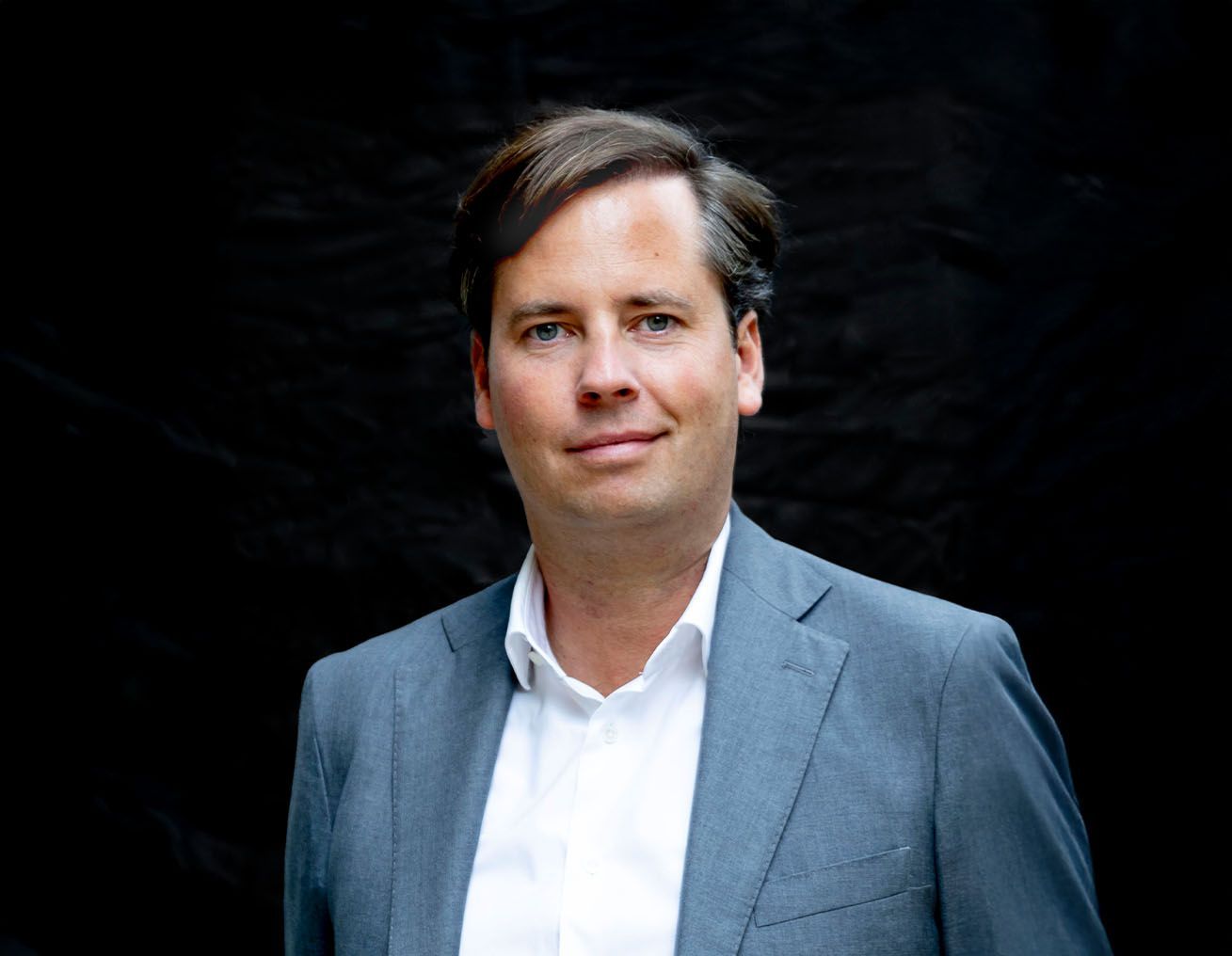 Een man in een pak en een wit overhemd staat voor een zwarte achtergrond.