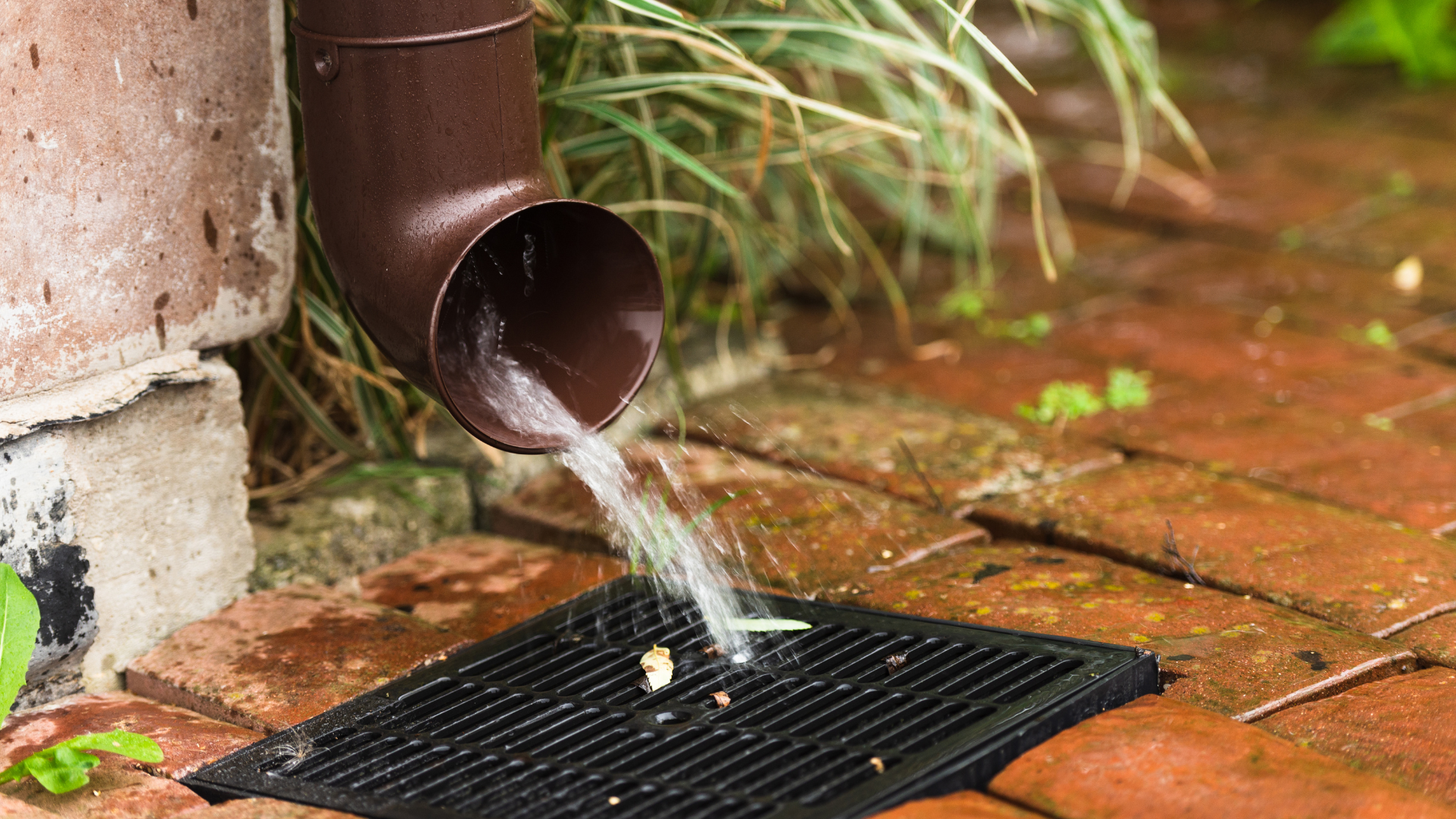 Brown downspout discharging water into a black drain on brick pavers.
