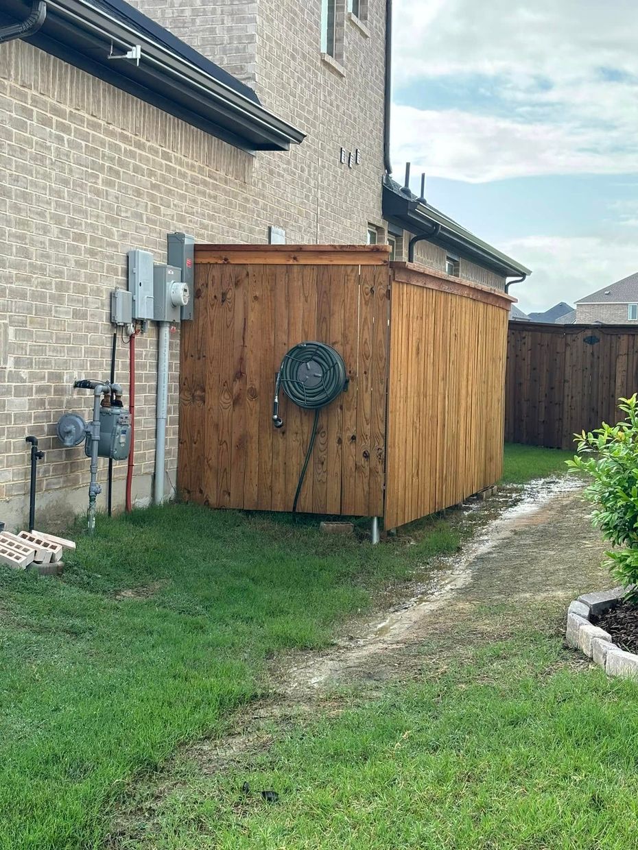 Wooden fence enclosure with a hose reel and electrical panel against a brick wall. Green grass and gravel path.