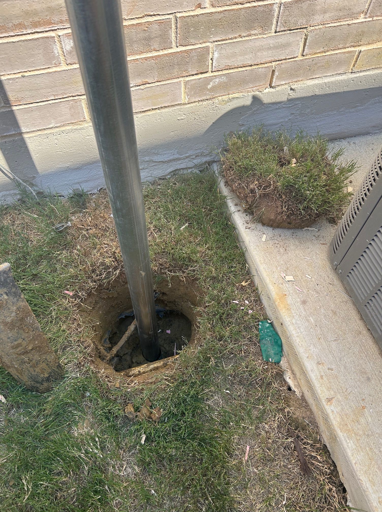 Pole set in ground with exposed hole, grass, and a brick wall.