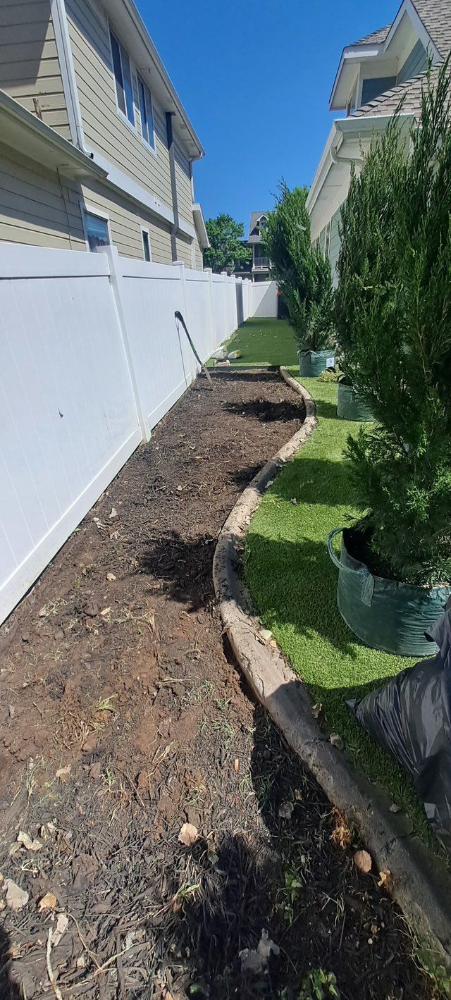 A narrow yard between two houses with a mulch bed, grass, and a white fence on a sunny day.
