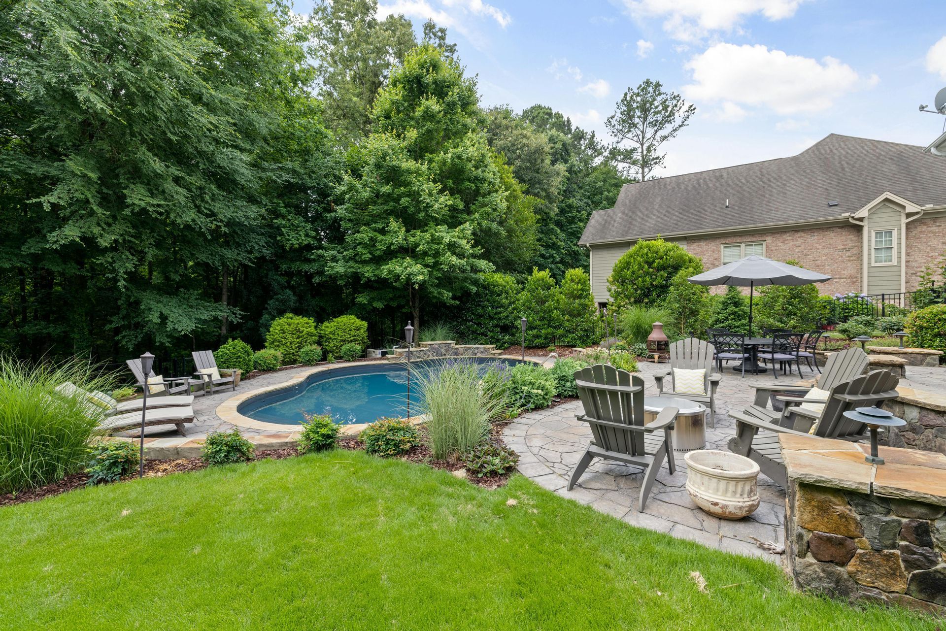 A backyard patio featuring a swimming pool, lounge chairs, dining furniture, and lush green trees near a brick house.