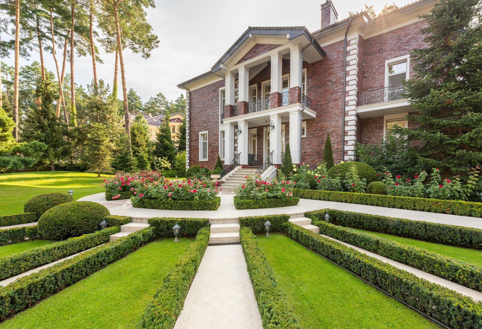 A formal brick mansion with white pillars set behind a symmetrical garden featuring manicured hedges and a stone path.