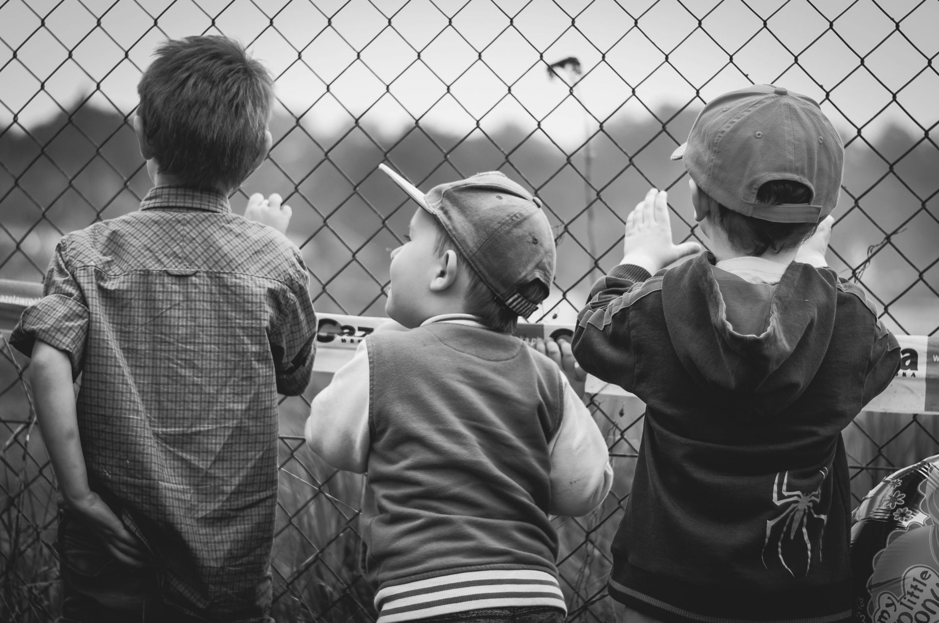 three children looking through a chainlink fence, presumably at a baseball field