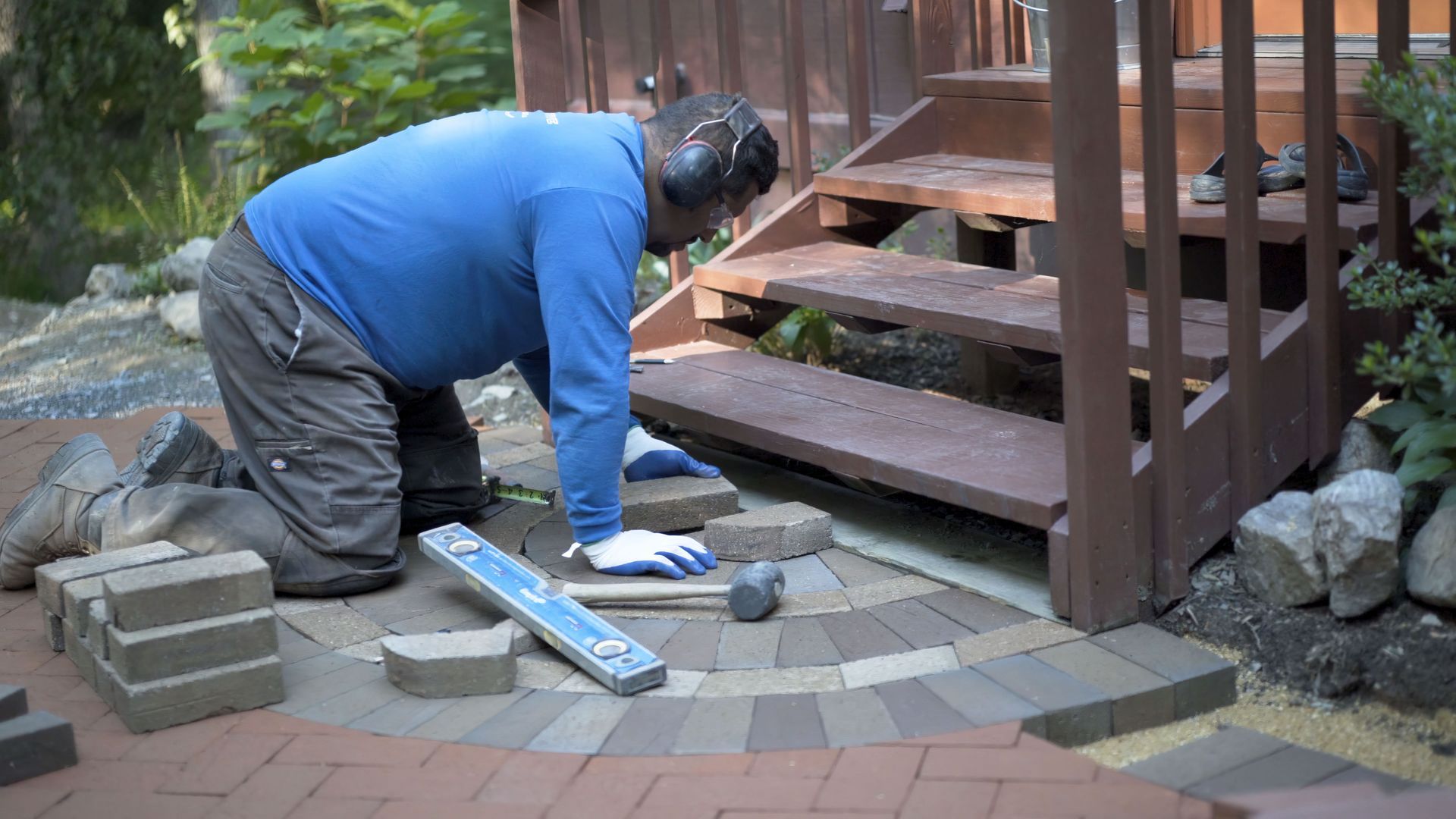 Man laying paving stones near wooden steps. He uses tools and wears ear protection outdoors.