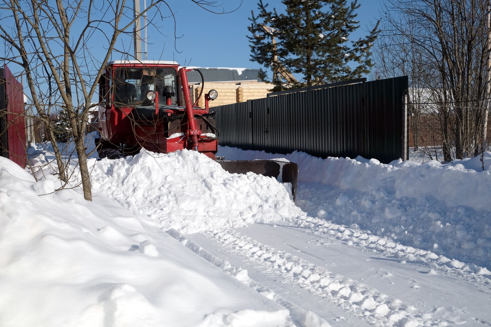 Red tractor plowing snow from a driveway, next to a black fence, on a sunny winter day.