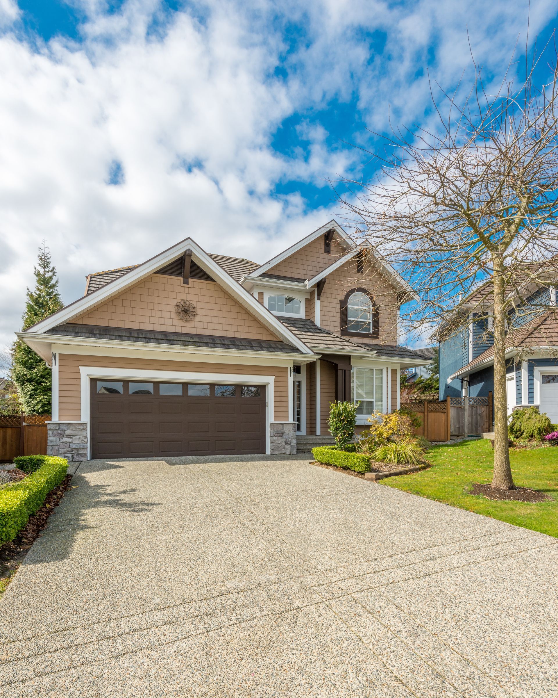 Beige and brown two-story house with a gravel driveway under a blue sky with clouds.