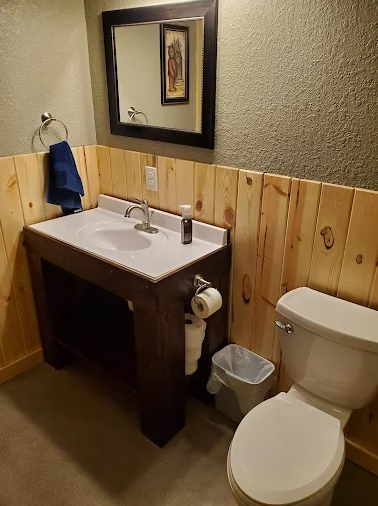 Small bathroom with white sink, toilet, mirror, wood wainscoting, and trash bin.