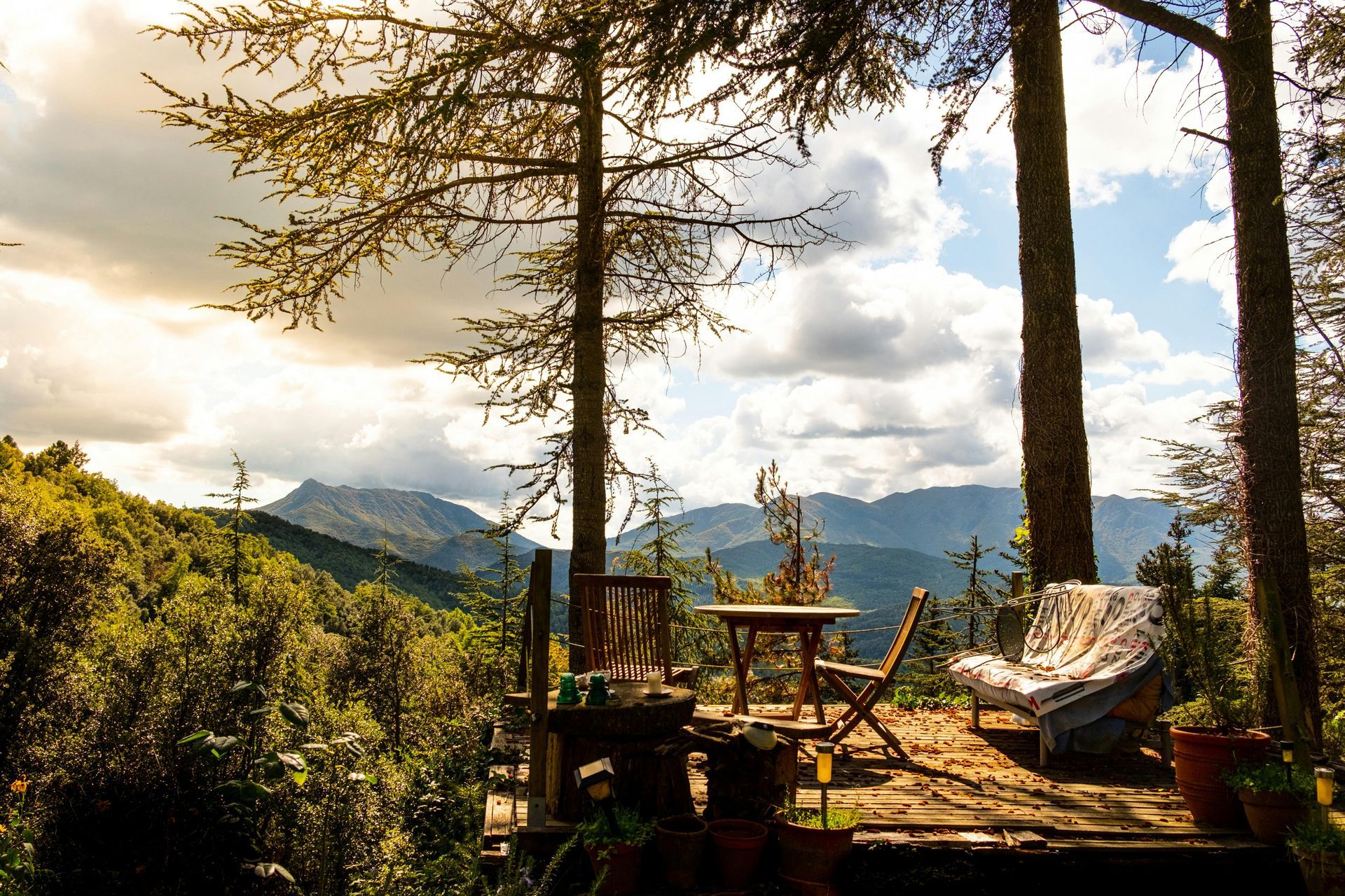 Mountain cabin deck with chairs under trees, overlooking a sunlit valley and cloudy sky